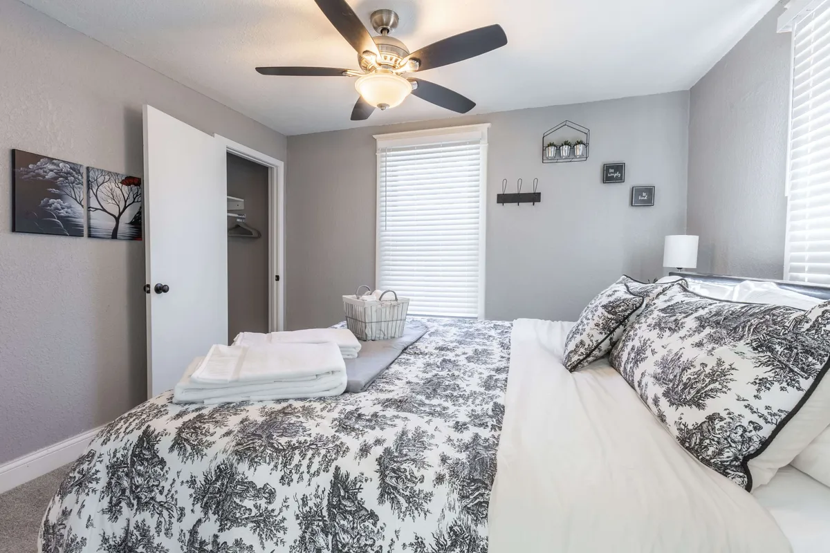 A serene bedroom scene with a bed dressed in patterned bedding, a ceiling fan overhead, a window with blinds letting in gentle light, and tasteful artwork on the wall. The ambiance is peaceful, perfect for relaxing or enjoying a quiet afternoon.
