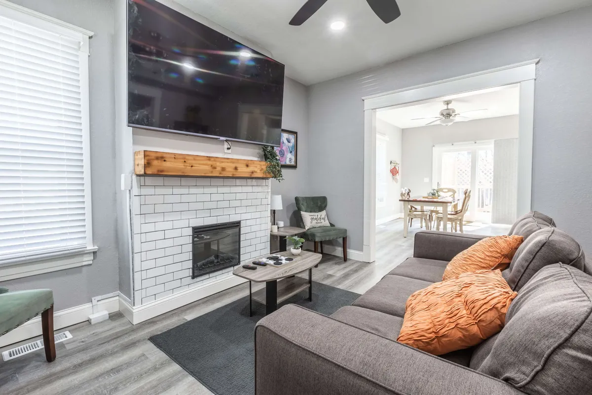 A spacious living room featuring a plush sofa with decorative pillows, a fireplace, a TV mounted on the wall, and green accent chairs. The open layout allows visibility into the adjacent dining area, creating a cohesive and inviting space for gatherings.