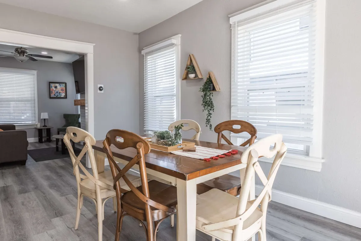 A spacious dining area with a large table surrounded by multiple chairs, complemented by decorative plants and ample natural light streaming in through large windows. The arrangement encourages both group gatherings and casual interactions.